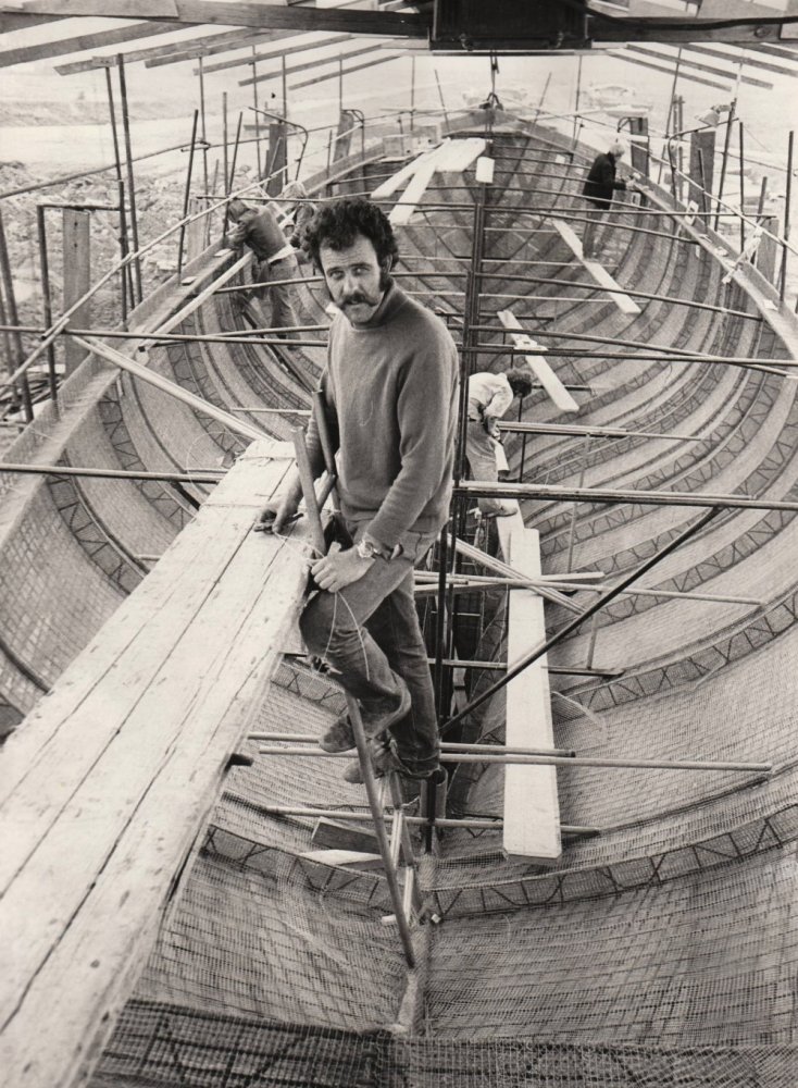 Man on scaffold inside large boat frame under construction, with people working in background.