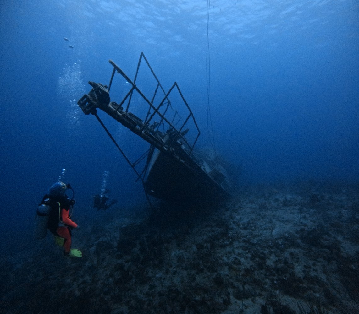 Two scuba divers explore a shipwreck on the ocean floor, surrounded by deep blue water.
