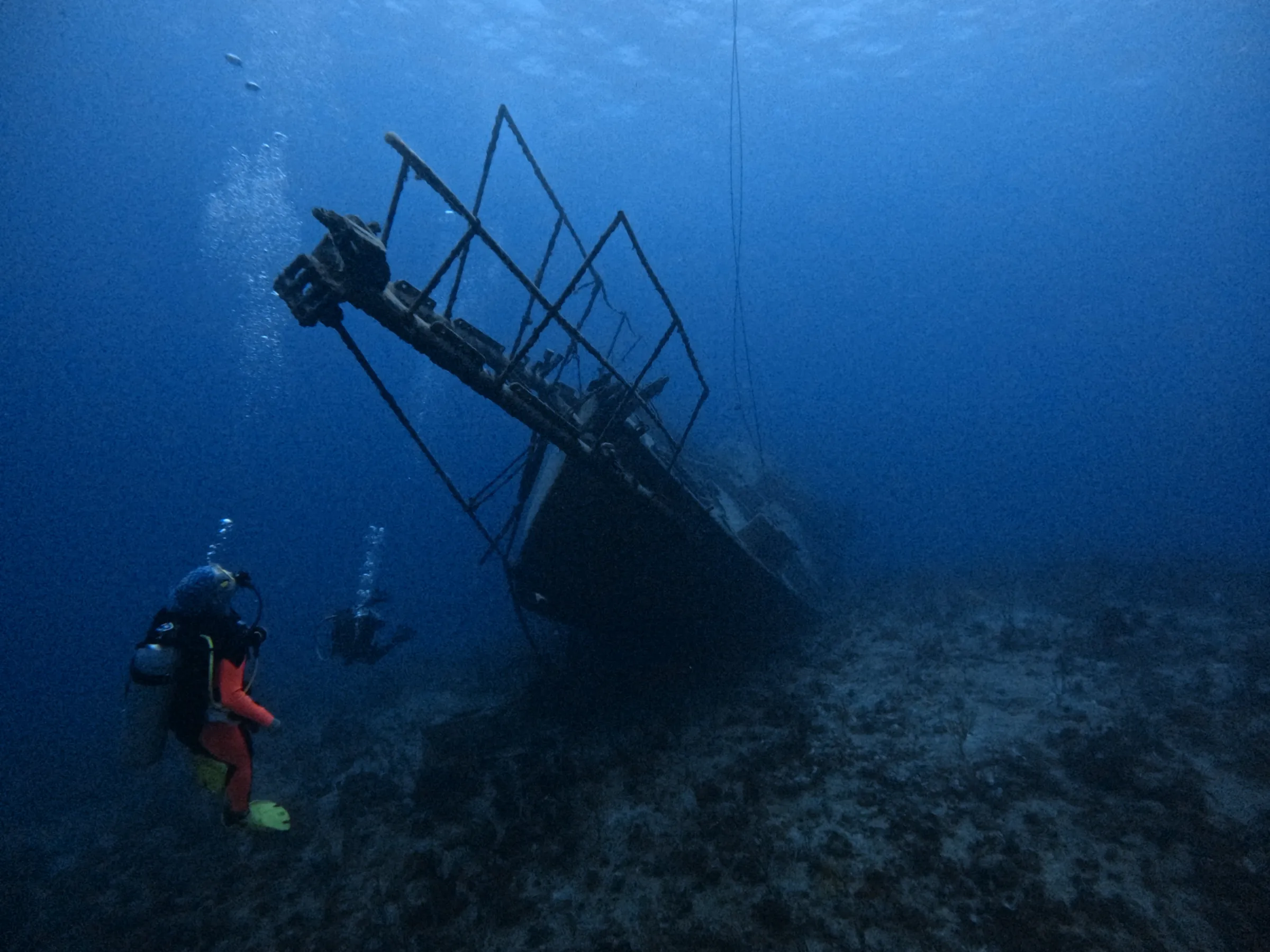 Two scuba divers explore a shipwreck on the ocean floor, surrounded by deep blue water.