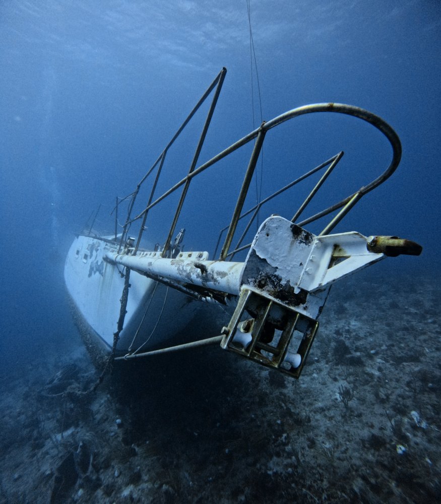 Underwater view of a sunken sailboat on the ocean floor, partially tilted and covered in marine growth.