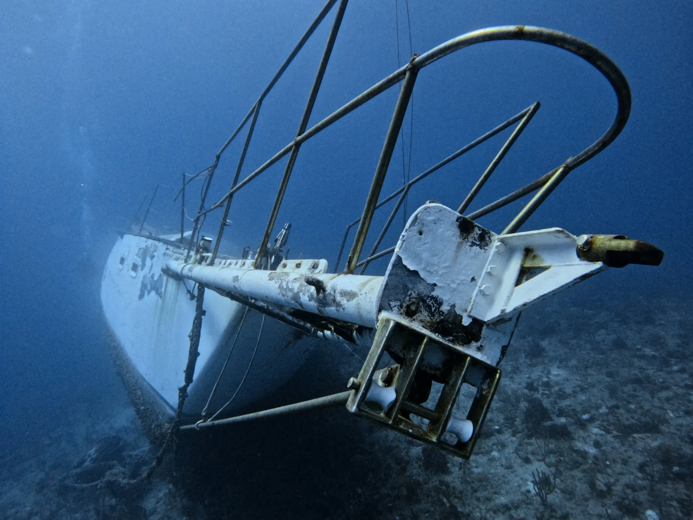 Underwater view of a sunken sailboat on the ocean floor, partially tilted and covered in marine growth.