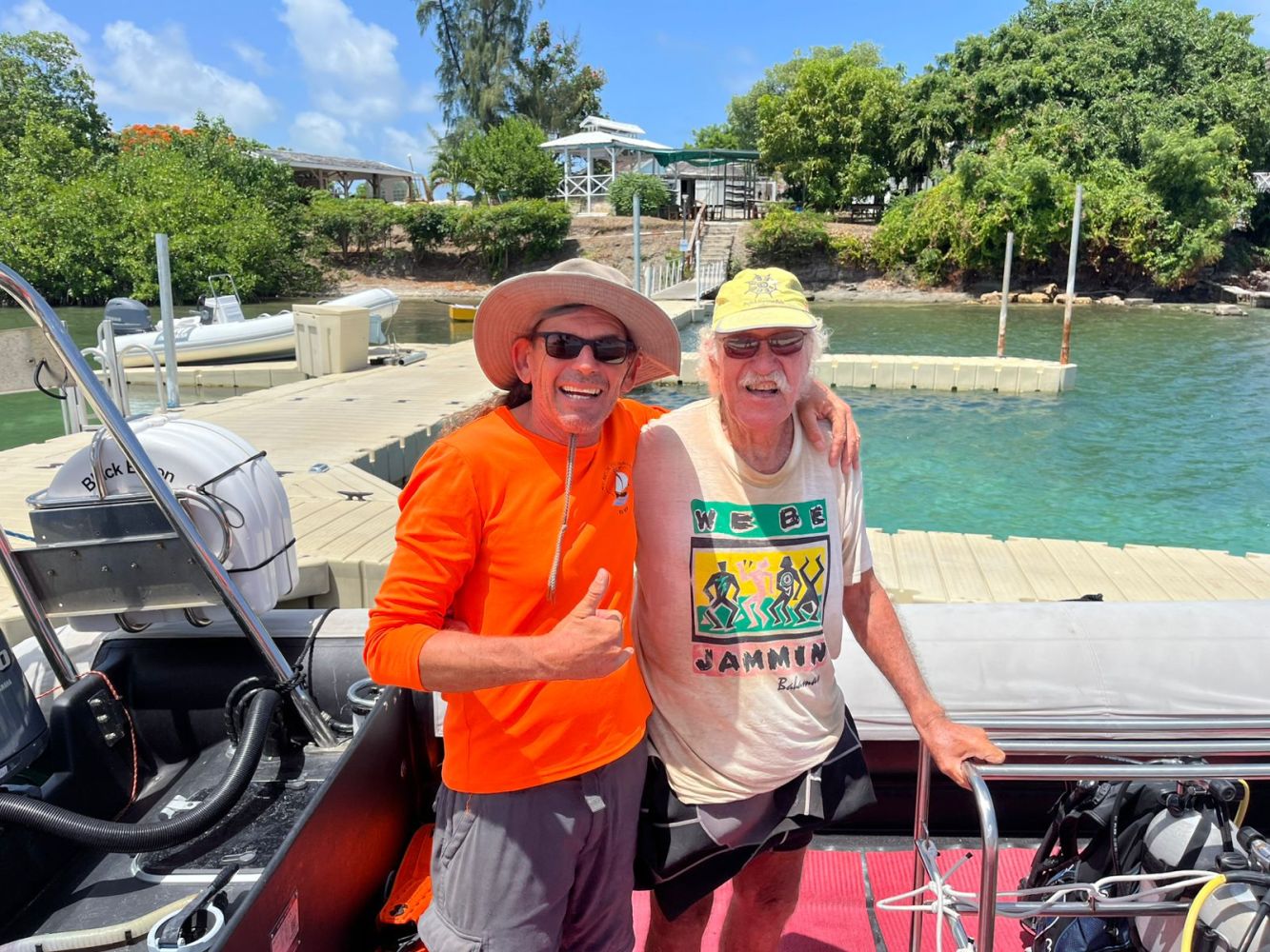 Two men smiling on a boat near a dock with trees and blue sky in the background.