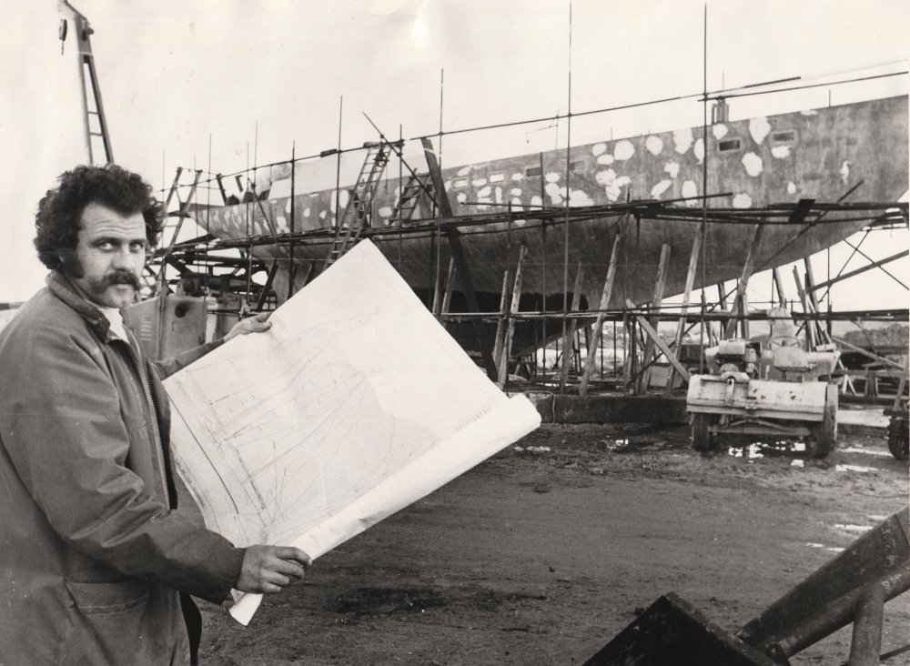 Man with mustache holds large blueprint in front of ship under construction with scaffolding.
